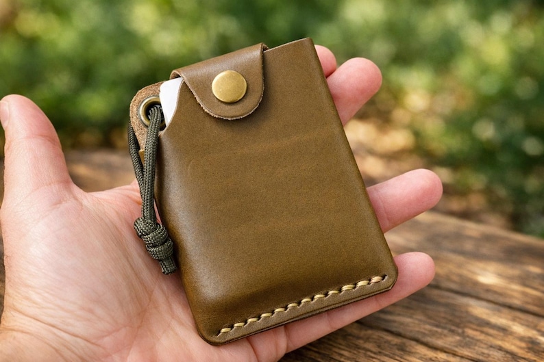 May include: An olive green leather card holder featuring a brass snap button closure, visible light-colored stitching along the bottom edge, and a matching olive green paracord lanyard attached through a reinforced metal eyelet on the side.