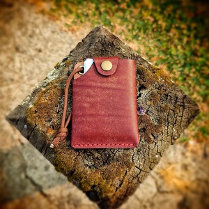May include: A rectangular reddish-brown leather card holder featuring a brass snap button closure, visible red stitching along the bottom edge, and a matching brown cord attached to a metal eyelet on the upper left corner, holding a small white card.
