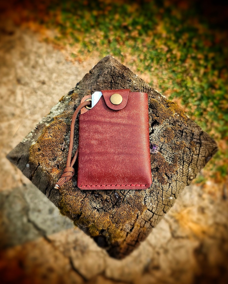 May include: A rectangular reddish-brown leather card holder featuring a brass snap button closure, visible red stitching along the bottom edge, and a matching brown cord attached to a metal eyelet on the upper left corner, holding a small white card.