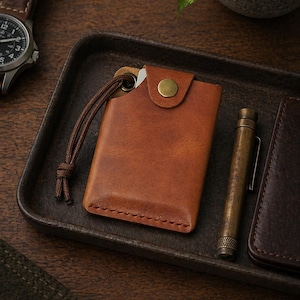 May include: A tan leather card holder featuring a brass snap button closure, visible stitching along the bottom edge, and a brown cord loop attached to a metal eyelet. The rectangular wallet is positioned next to a metallic pen and a watch.