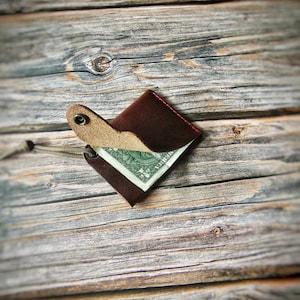 May include: A dark brown leather wallet with a single snap button closure and red stitching. The wallet is partially open, revealing a one-dollar bill tucked inside. A thin, light-colored cord is attached to a metal eyelet on the side of the wallet.