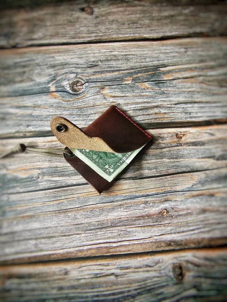 May include: A dark brown leather wallet with a single snap button closure and red stitching. The wallet is partially open, revealing a one-dollar bill tucked inside. A thin, light-colored cord is attached to a metal eyelet on the side of the wallet.
