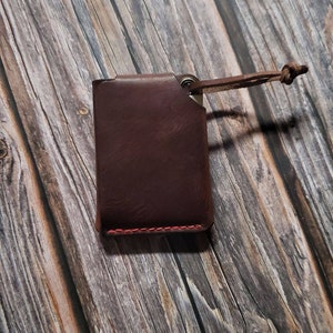 May include: A dark brown leather card holder featuring a rectangular shape with visible red stitching along the bottom edge. A small metal eyelet is attached to the top right corner, connected to a thin leather cord with a knotted end.