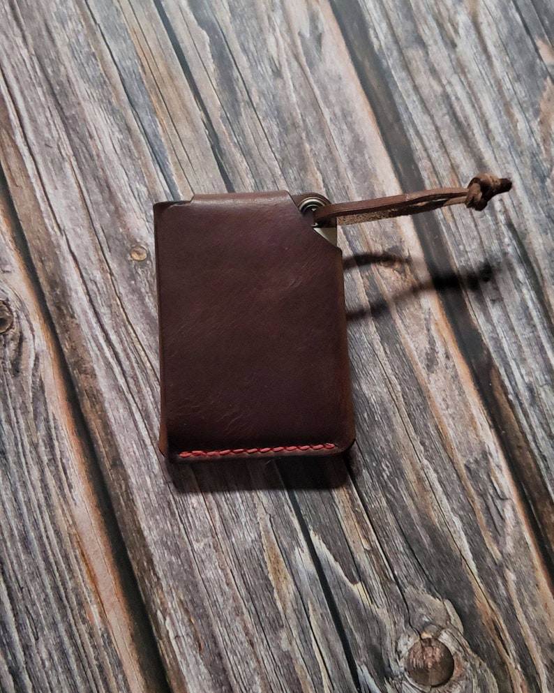 May include: A dark brown leather card holder featuring a rectangular shape with visible red stitching along the bottom edge. A small metal eyelet is attached to the top right corner, connected to a thin leather cord with a knotted end.