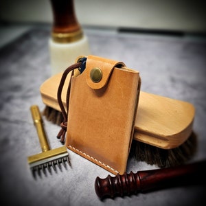 May include: A tan leather card holder featuring white stitching, a brass snap button closure, and a brown cord pull tab. The rectangular wallet is constructed from smooth, light brown leather with visible grain and a secure flap design.