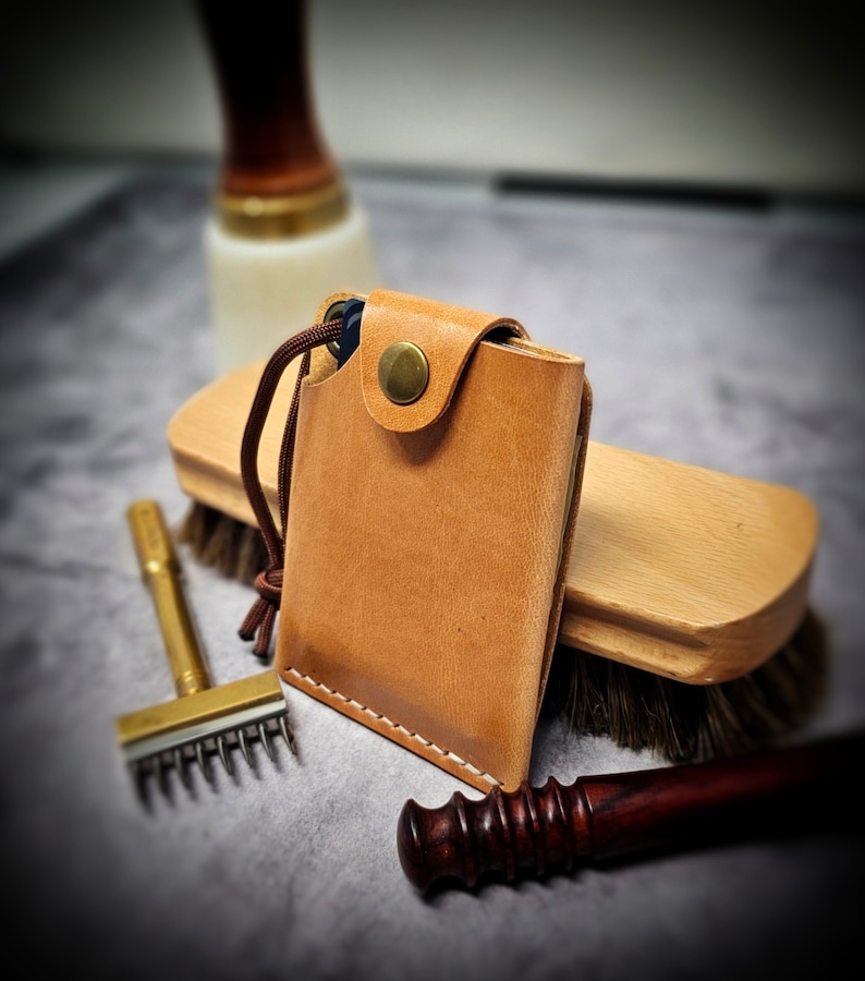 May include: A tan leather card holder featuring white stitching, a brass snap button closure, and a brown cord pull tab. The rectangular wallet is constructed from smooth, light brown leather with visible grain and a secure flap design.