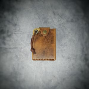 May include: A tan leather wallet featuring a distressed texture, a brass-colored snap button closure, and a brown cord attached to a metal grommet on the top left corner. The wallet has visible stitching along the bottom edge and a rectangular shape.