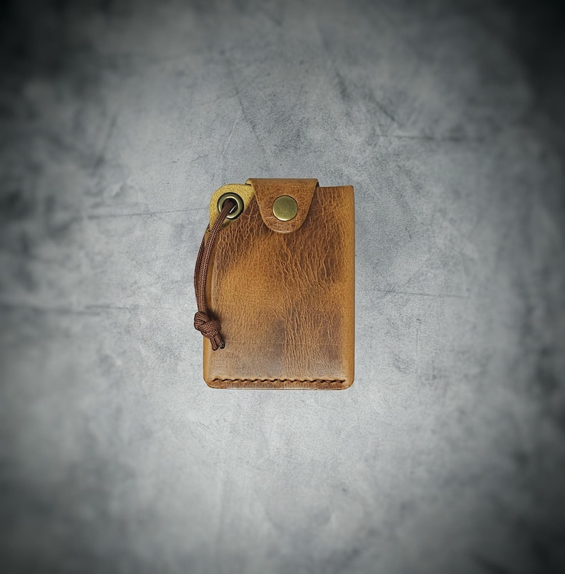 May include: A tan leather wallet featuring a distressed texture, a brass-colored snap button closure, and a brown cord attached to a metal grommet on the top left corner. The wallet has visible stitching along the bottom edge and a rectangular shape.