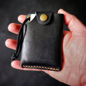 May include: A black leather card holder featuring a brass snap button closure, a black cord lanyard attached through a metal eyelet, and visible tan stitching along the bottom edge. The rectangular wallet has a smooth finish and a flap top.