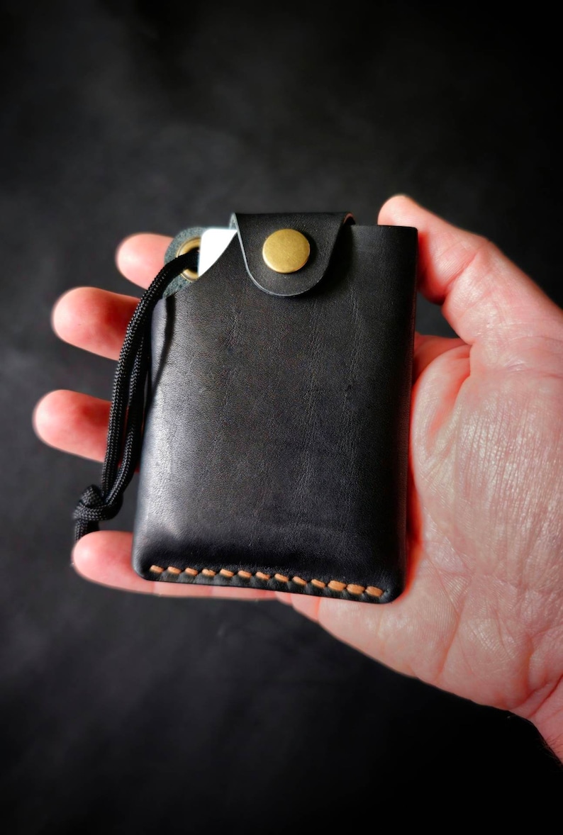 May include: A black leather card holder featuring a brass snap button closure, a black cord lanyard attached through a metal eyelet, and visible tan stitching along the bottom edge. The rectangular wallet has a smooth finish and a flap top.