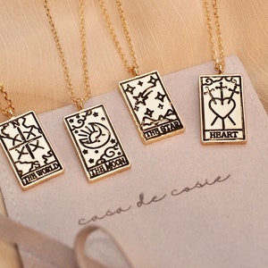 May include: Four gold-colored tarot card pendant necklaces. Each rectangular pendant features a black design and text, including "THE WORLD," "THE MOON," "THE STAR," and "HEART." The necklaces rest on a light-colored fabric.