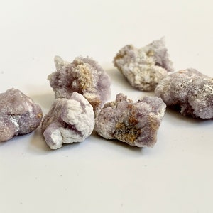 May include: A group of six small, rough, purple and white amethyst crystals. The crystals are clustered together on a white surface.