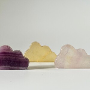 May include: Three cloud-shaped decorative stones. One is a deep purple with lighter purple streaks, the second is a translucent yellow, and the third is a pale pink. The stones are smooth and sit on a white surface.
