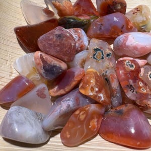 May include: A collection of polished gemstones in shades of red, orange, pink, and white. The stones vary in size and shape, with smooth, rounded surfaces. Some stones have unique patterns and inclusions, and are displayed in a wooden bowl.