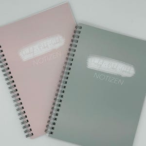 Könnte beinhalten: Zwei spiralgebundene Notizbücher, eines in zartem Rosa und das andere in gedämpftem Grün. Jedes Notizbuch trägt den Text "Predigt Bibel Gebet" und "NOTIZEN" in weißer, handschriftlicher Schrift. Die Notizbücher liegen auf einer weißen Oberfläche.