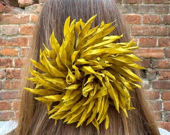 May include: A large, yellow-green hair accessory, resembling a stylized flower, is attached to the back of a person's head. The accessory is made of many leaf-like elements. The person has long, brown hair, and the background is a brick wall.