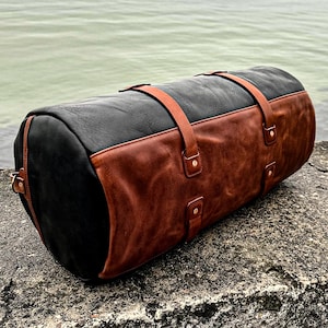 May include: A cylindrical duffel bag constructed from black leather with a large brown leather panel covering the front section. The bag features two brown leather straps that wrap around the body, secured with metal rivets at the attachment points.
