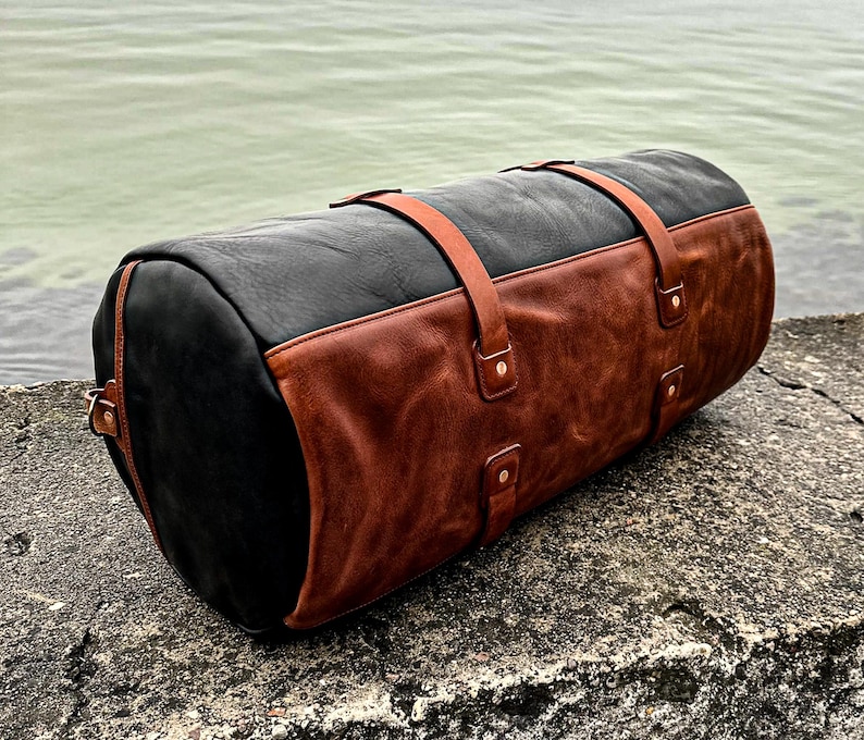 May include: A cylindrical duffel bag constructed from black leather with a large brown leather panel covering the front section. The bag features two brown leather straps that wrap around the body, secured with metal rivets at the attachment points.