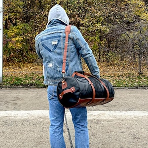 May include: A cylindrical duffel bag featuring a black main body with brown leather accents, including two encircling straps, a side handle, and an adjustable shoulder strap with metal hardware, worn across the back over a denim jacket and jeans.