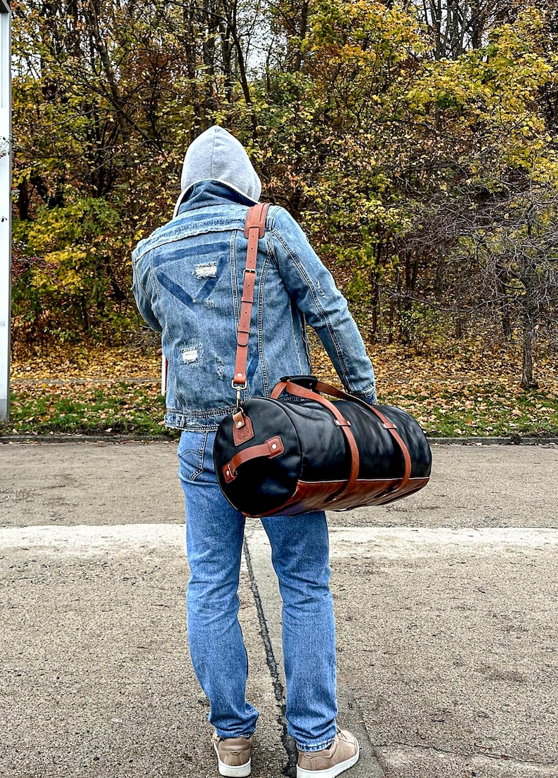 May include: A cylindrical duffel bag featuring a black main body with brown leather accents, including two encircling straps, a side handle, and an adjustable shoulder strap with metal hardware, worn across the back over a denim jacket and jeans.