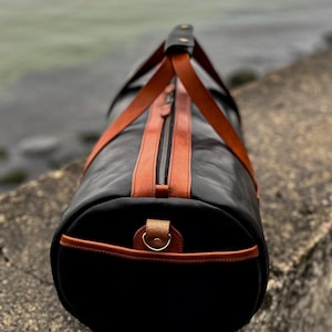 May include: A cylindrical duffel bag crafted from black leather with contrasting tan leather accents. The bag features a central zipper closure, two tan leather handles that meet at a top grip, and a metal D-ring attached to the circular end panel.