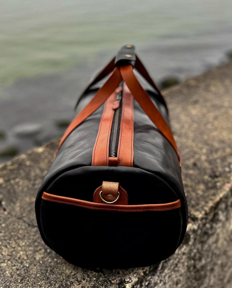 May include: A cylindrical duffel bag crafted from black leather with contrasting tan leather accents. The bag features a central zipper closure, two tan leather handles that meet at a top grip, and a metal D-ring attached to the circular end panel.
