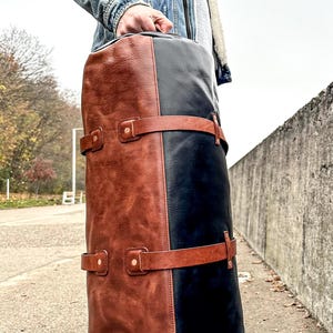 May include: A cylindrical duffel bag constructed from two-tone leather, featuring a rich brown panel on one side and a deep black panel on the other. Two horizontal brown leather straps with metallic rivets wrap around the exterior body.
