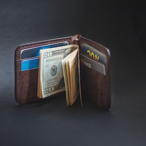 May include: A brown leather wallet with a money clip, holding several twenty dollar bills and two credit cards. One card has the text "MAXI" and the other has the text "200%.