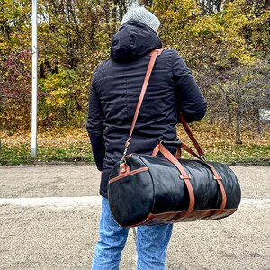 May include: A cylindrical duffel bag featuring a black main body with brown leather trim, accents, and a long adjustable shoulder strap. The bag includes two top handles and is worn across the body, showcasing a structured design with metal hardware.