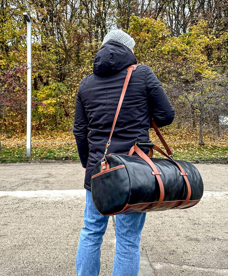May include: A cylindrical duffel bag featuring a black main body with brown leather trim, accents, and a long adjustable shoulder strap. The bag includes two top handles and is worn across the body, showcasing a structured design with metal hardware.