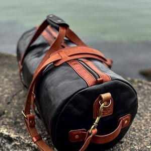 May include: A cylindrical duffel bag crafted from black leather with contrasting tan leather accents, including top handles, a shoulder strap, and trim. The bag features a central zipper closure, brass-toned metal hardware, and side handles.