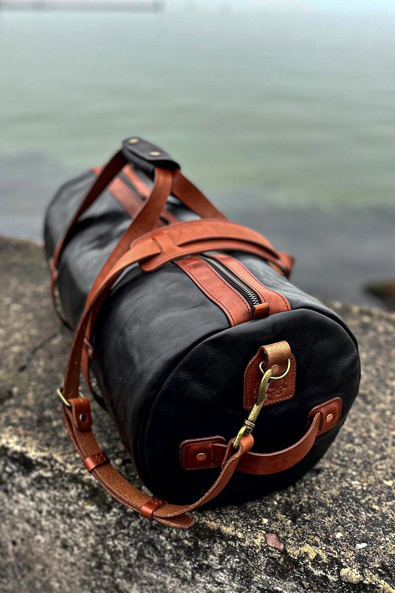 May include: A cylindrical duffel bag crafted from black leather with contrasting tan leather accents, including top handles, a shoulder strap, and trim. The bag features a central zipper closure, brass-toned metal hardware, and side handles.