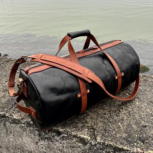 May include: A black leather duffel bag featuring tan leather straps, a top handle with a black grip, and a detachable shoulder strap. The bag includes metal hardware, side handles, and a zippered closure, showcasing a structured cylindrical design.