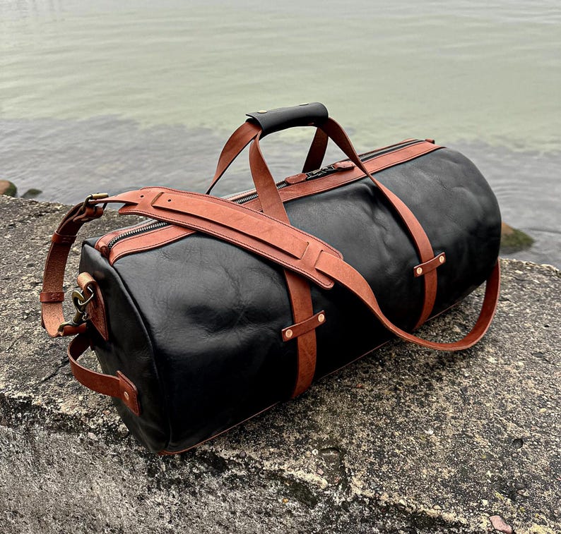 May include: A black leather duffel bag featuring tan leather straps, a top handle with a black grip, and a detachable shoulder strap. The bag includes metal hardware, side handles, and a zippered closure, showcasing a structured cylindrical design.