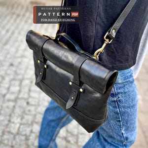 Può includere: Una borsa a tracolla in pelle nera con patta ripiegabile, finiture in metallo color ottone e tracolla regolabile. La borsa presenta cinghie in pelle verticali con rivetti in ottone. Il testo recita: MUSAK PATTERNS PATTERN PDF FOR HAND STITCHING.