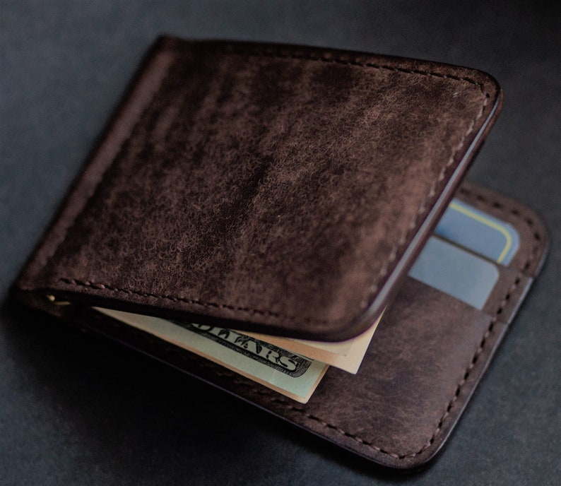 May include: A brown leather wallet with a money clip and a slot for credit cards. The wallet is open and there is a dollar bill inside.