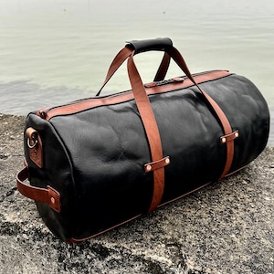 May include: A cylindrical duffel bag crafted from black leather with contrasting tan leather straps, handles, and trim. The bag features a top zipper closure, a side handle, and metallic rivets securing the straps to the main body of the bag.
