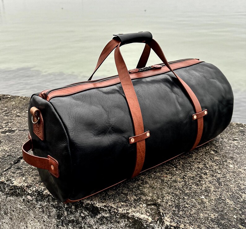 May include: A cylindrical duffel bag crafted from black leather with contrasting tan leather straps, handles, and trim. The bag features a top zipper closure, a side handle, and metallic rivets securing the straps to the main body of the bag.
