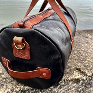 May include: A cylindrical duffel bag crafted from black leather with tan leather accents. It features two long top handles, a side handle, a metal zipper closure, and a debossed logo patch on the side. The bag is constructed with brass-toned hardware.