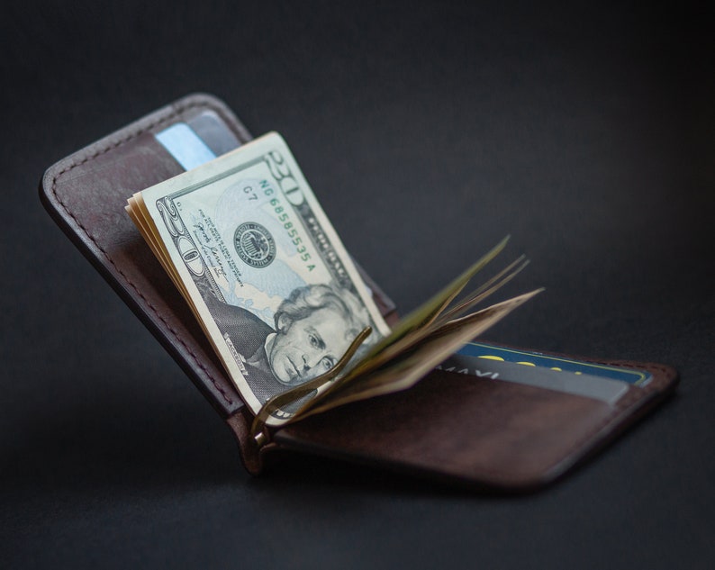 May include: A brown leather wallet with a money clip holding a stack of US dollar bills. The wallet has a credit card slot and a flap that closes with a snap.