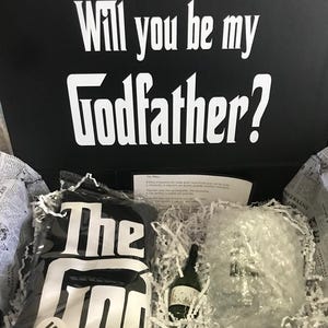 Könnte beinhalten: Schwarze Geschenkbox mit dem weißen Text "Will you be my Godfather?". Darin ein schwarzes T-Shirt mit "The God" in Weiß, eine kleine Flasche und ein verpacktes Glas. Die Box ist mit Zeitungspapier ausgekleidet.