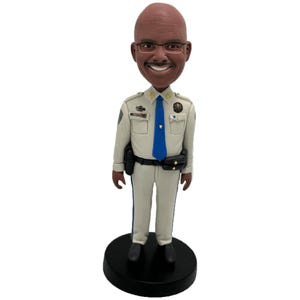 May include: A bobblehead figurine of a smiling man wearing glasses and a police uniform. The uniform is light tan with a blue tie and black belt. The figurine is standing on a black circular base.