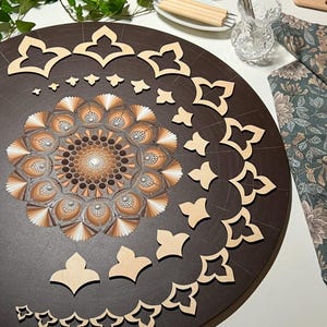 Puede incluir: Un tablero redondo de madera marr&oacute;n con un dise&ntilde;o de mandala pintado en tonos marrones y blancos. El tablero est&aacute; rodeado de plantillas de madera en forma de tr&eacute;bol de cuatro hojas.