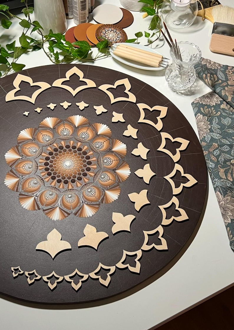 Puede incluir: Un tablero redondo de madera marr&oacute;n con un dise&ntilde;o de mandala pintado en tonos marrones y blancos. El tablero est&aacute; rodeado de plantillas de madera en forma de tr&eacute;bol de cuatro hojas.
