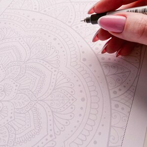 Puede incluir: Primer plano de una mano que utiliza un bol&iacute;grafo negro para delinear un dise&ntilde;o de mandala detallado en una hoja de papel blanca. El mandala es un patr&oacute;n circular con detalles intrincados y un color gris claro.