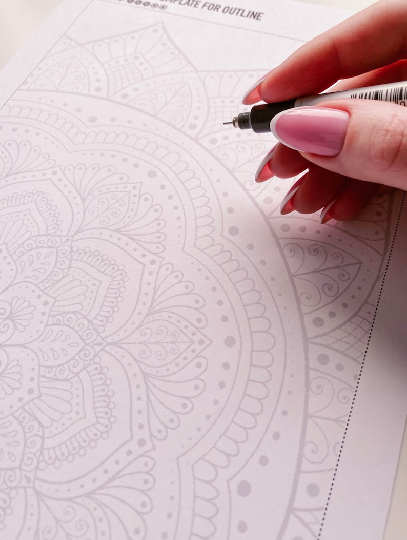 Puede incluir: Primer plano de una mano que utiliza un bol&iacute;grafo negro para delinear un dise&ntilde;o de mandala detallado en una hoja de papel blanca. El mandala es un patr&oacute;n circular con detalles intrincados y un color gris claro.