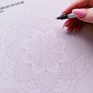 Puede incluir: Una plantilla de papel blanco con el texto "Template for Outline" en la parte superior. Un dise&ntilde;o de mandala detallado est&aacute; impreso en gris claro en el centro de la p&aacute;gina. Una mano sostiene un bol&iacute;grafo marcador negro y est&aacute; a punto de trazar el contorno del mandala.