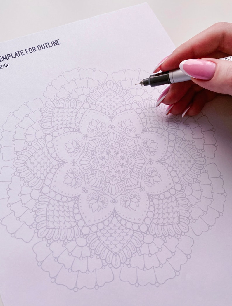 Puede incluir: Una plantilla de papel blanco con el texto "Template for Outline" en la parte superior. Un dise&ntilde;o de mandala detallado est&aacute; impreso en gris claro en el centro de la p&aacute;gina. Una mano sostiene un bol&iacute;grafo marcador negro y est&aacute; a punto de trazar el contorno del mandala.