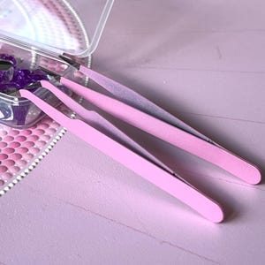 May include: Two pairs of pink tweezers, one pair is slightly larger than the other, on a pink surface.