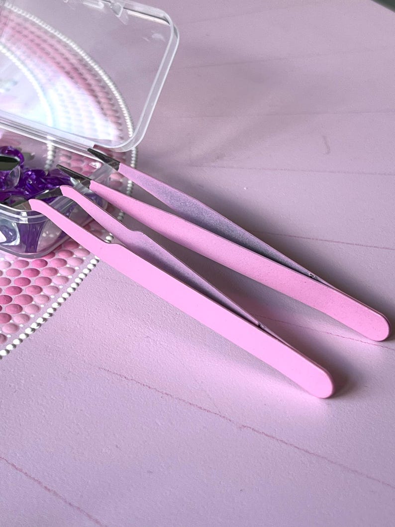 May include: Two pairs of pink tweezers, one pair is slightly larger than the other, on a pink surface.